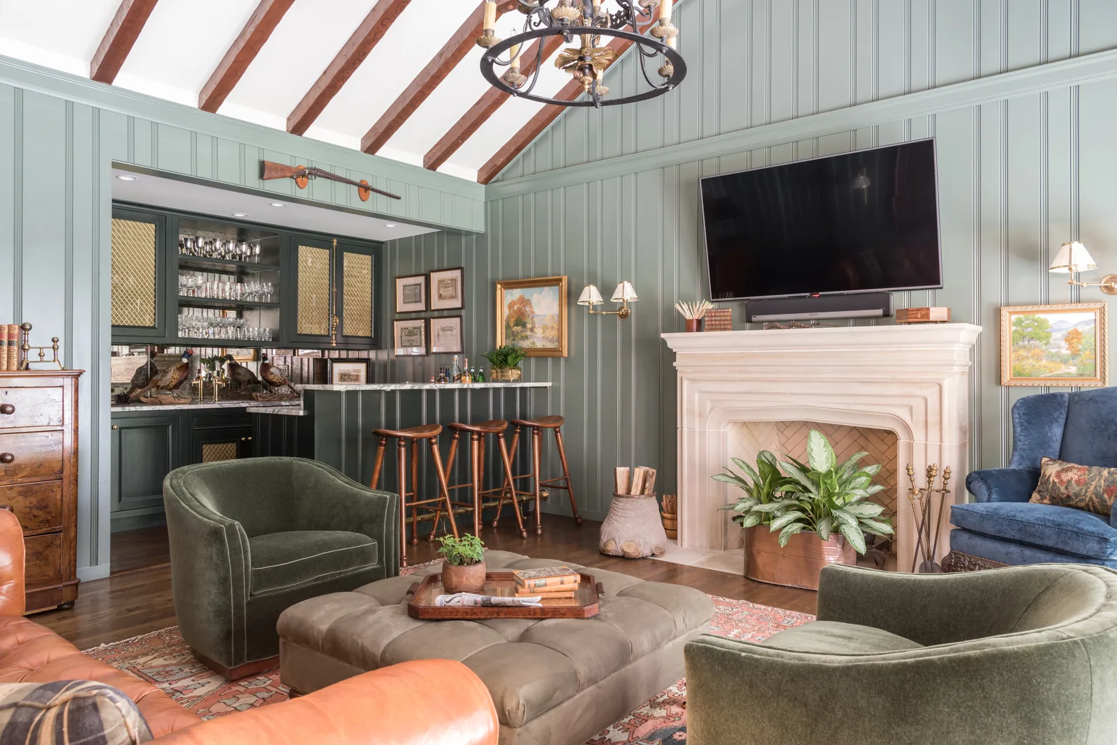 Living room under a vaulted beamed ceiling with sage panelled walls, a limestone fireplace, green velvet swivel chairs and a leather ottoman