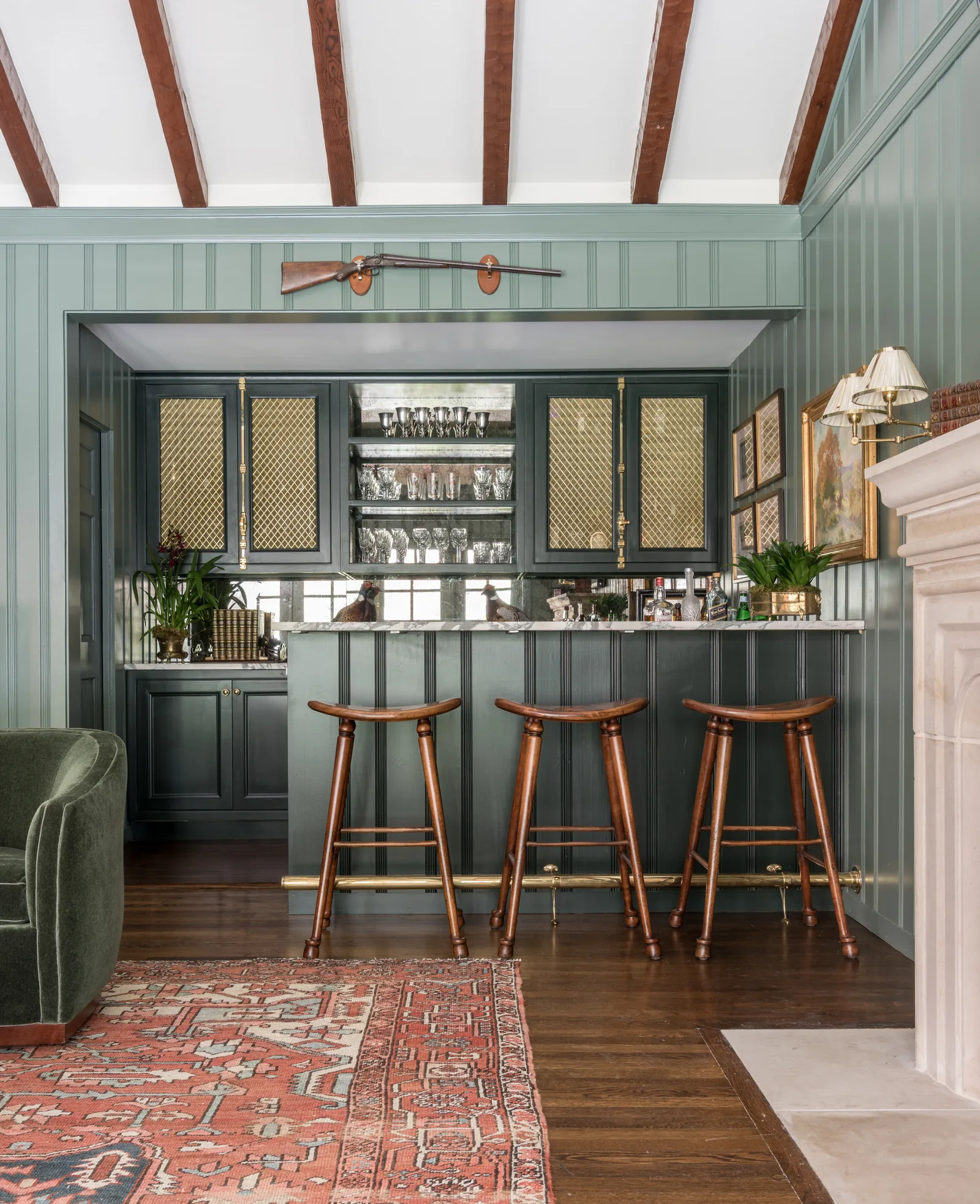 Sage green panelled bar with brass mesh cabinet doors, three wooden saddle stools and an antique rifle mounted above